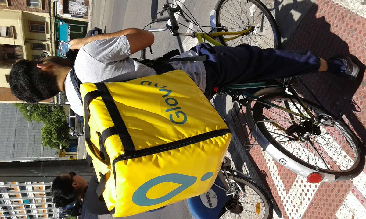 Repartidor de Glovo en bicicleta urbana con la mochila amarilla característica de la plataforma, fotografiado desde atrás