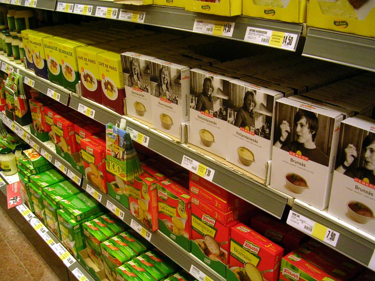 Lineal de supermercado con productos de marca blanca y marcas comerciales conviviendo en estantería, con etiquetas amarillas de precio