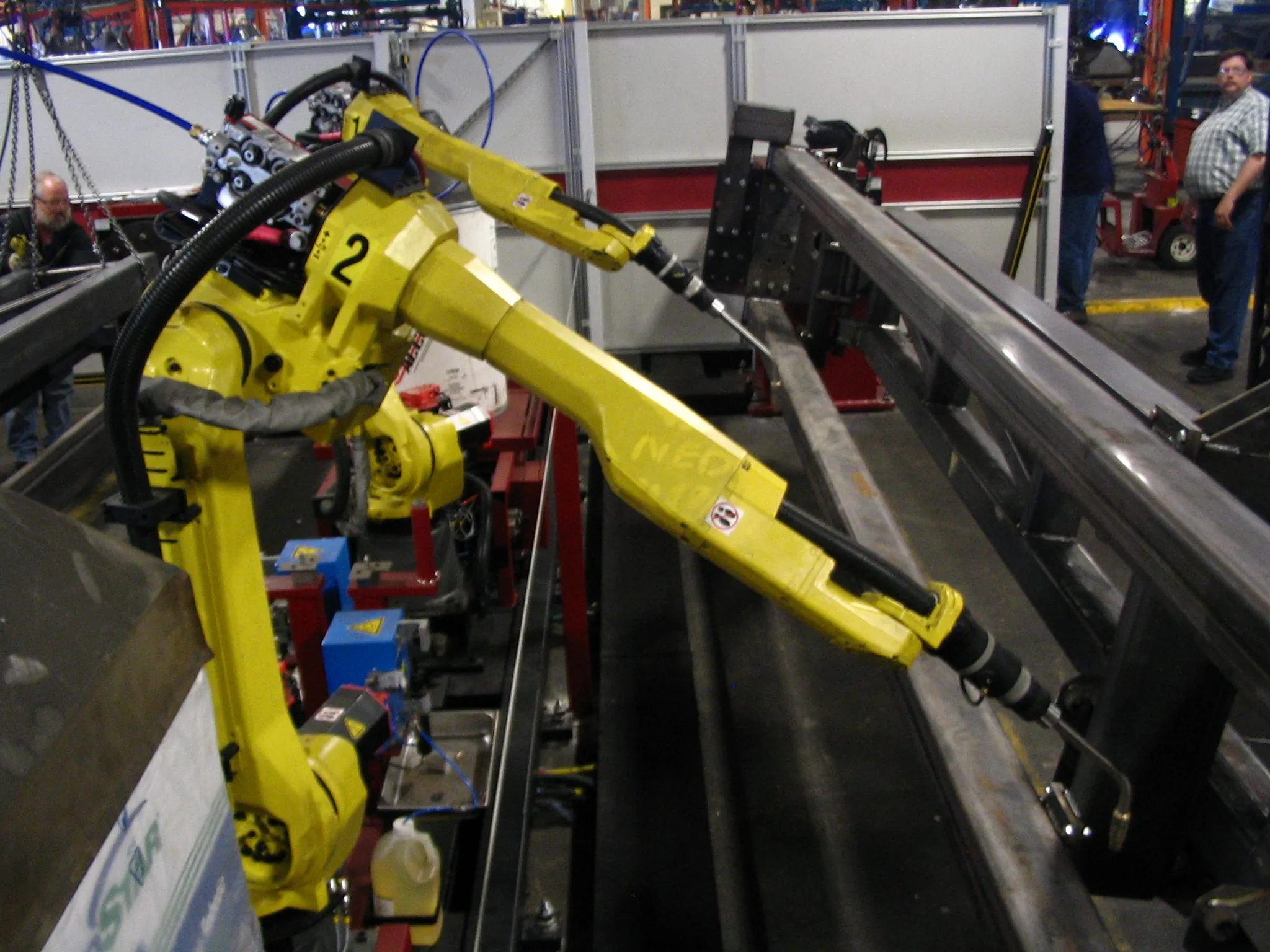 Brazo robótico FANUC amarillo de seis ejes soldando en línea industrial, con operario humano supervisando al fondo