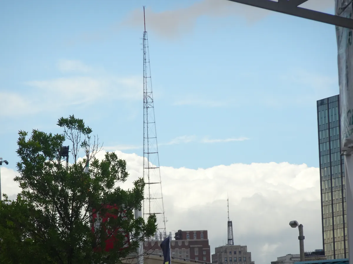 Torre antena de telecomunicaciones sobre azotea en entorno urbano, con árboles, edificio acristalado contemporáneo y cielo nublado al fondo