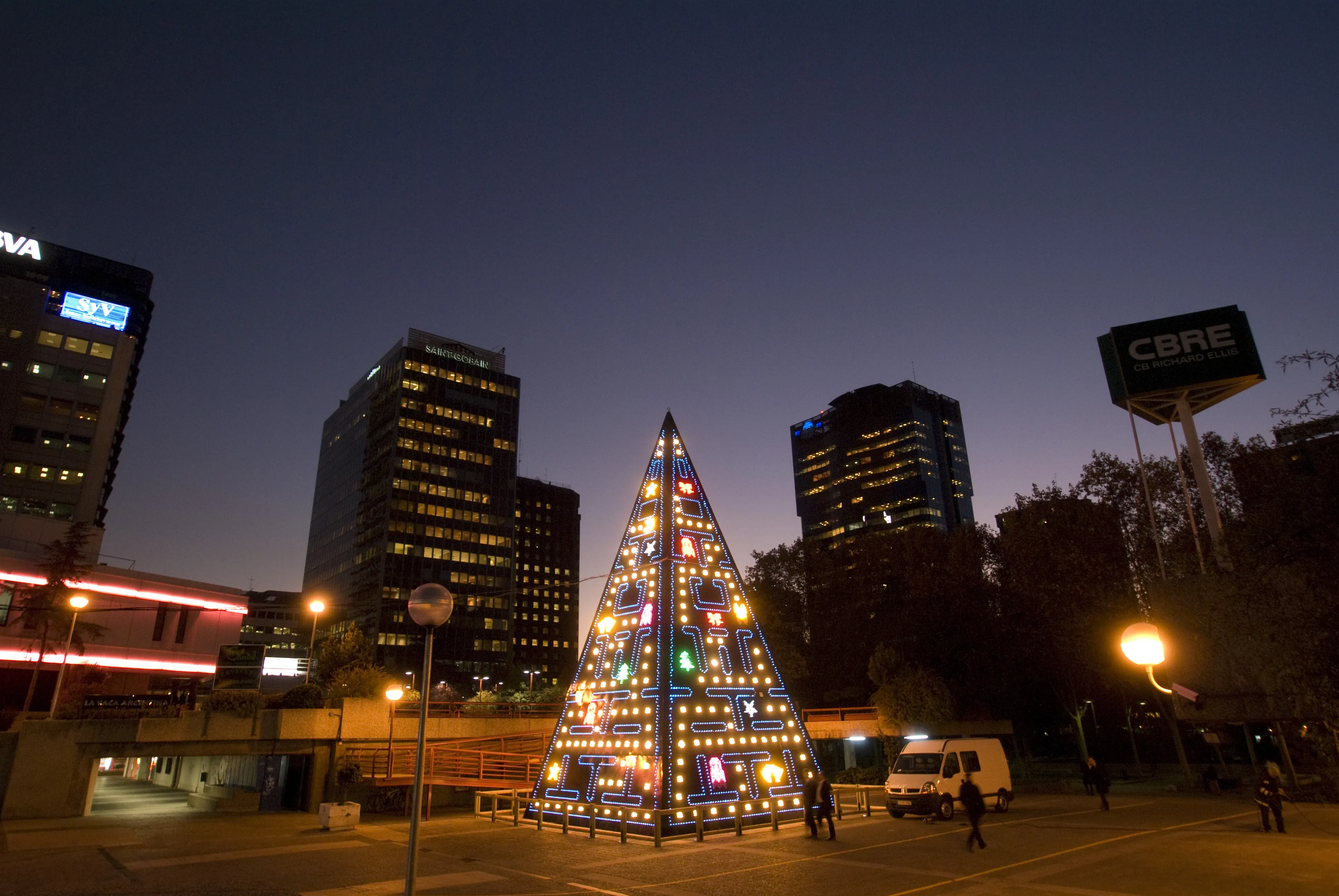 AZCA en Madrid al anochecer, con torres BBVA y CBRE iluminadas y un árbol-pirámide de luces de colores en primer plano