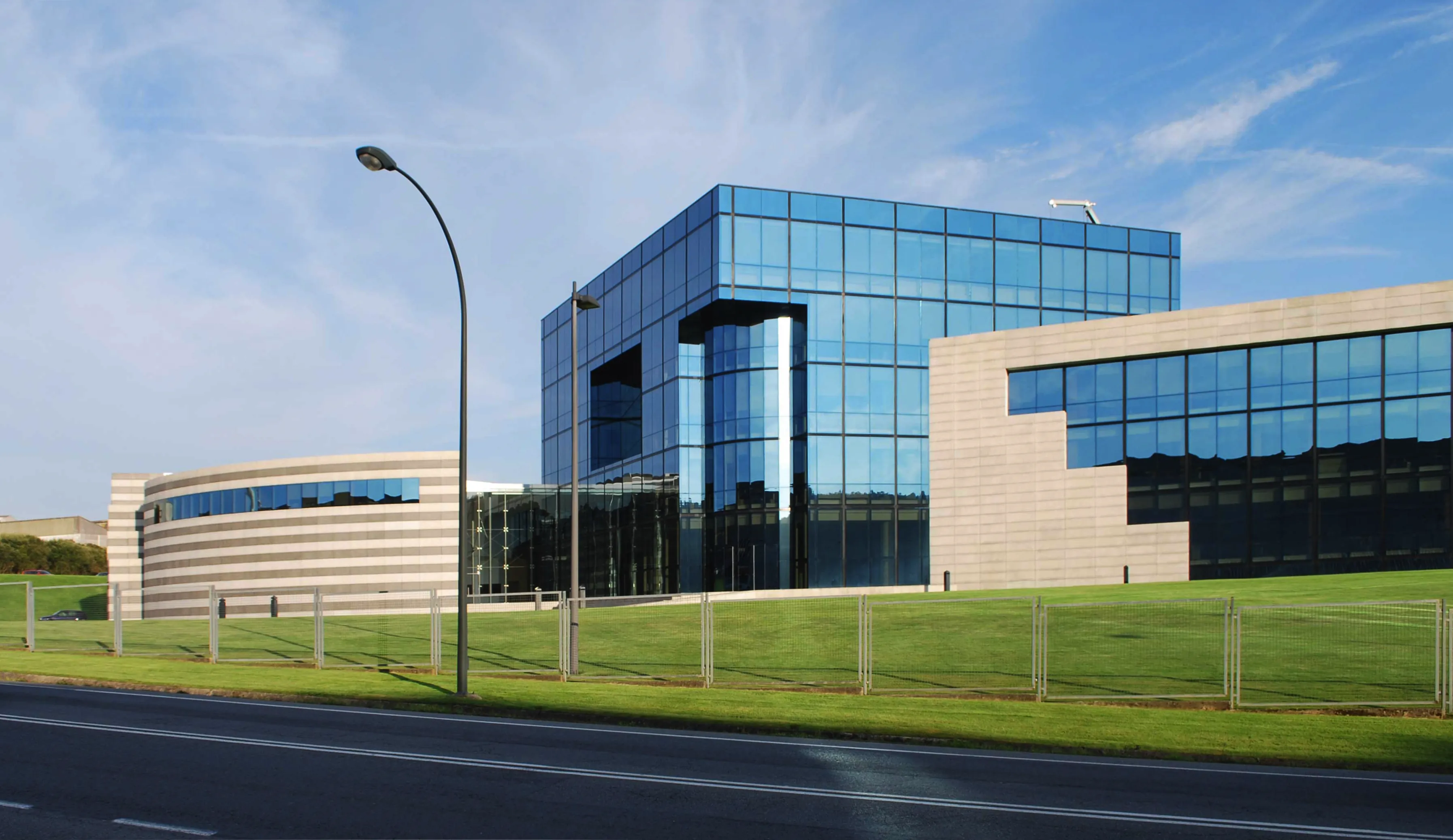 Sede de Inditex en Arteixo (A Coruña), edificio de oficinas de cristal azulado con módulo bajo de hormigón, sobre césped y junto a carretera